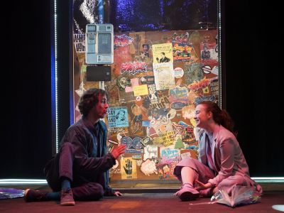Two actors sit on the floor of the set in front of a phone booth covered in posters and stickers